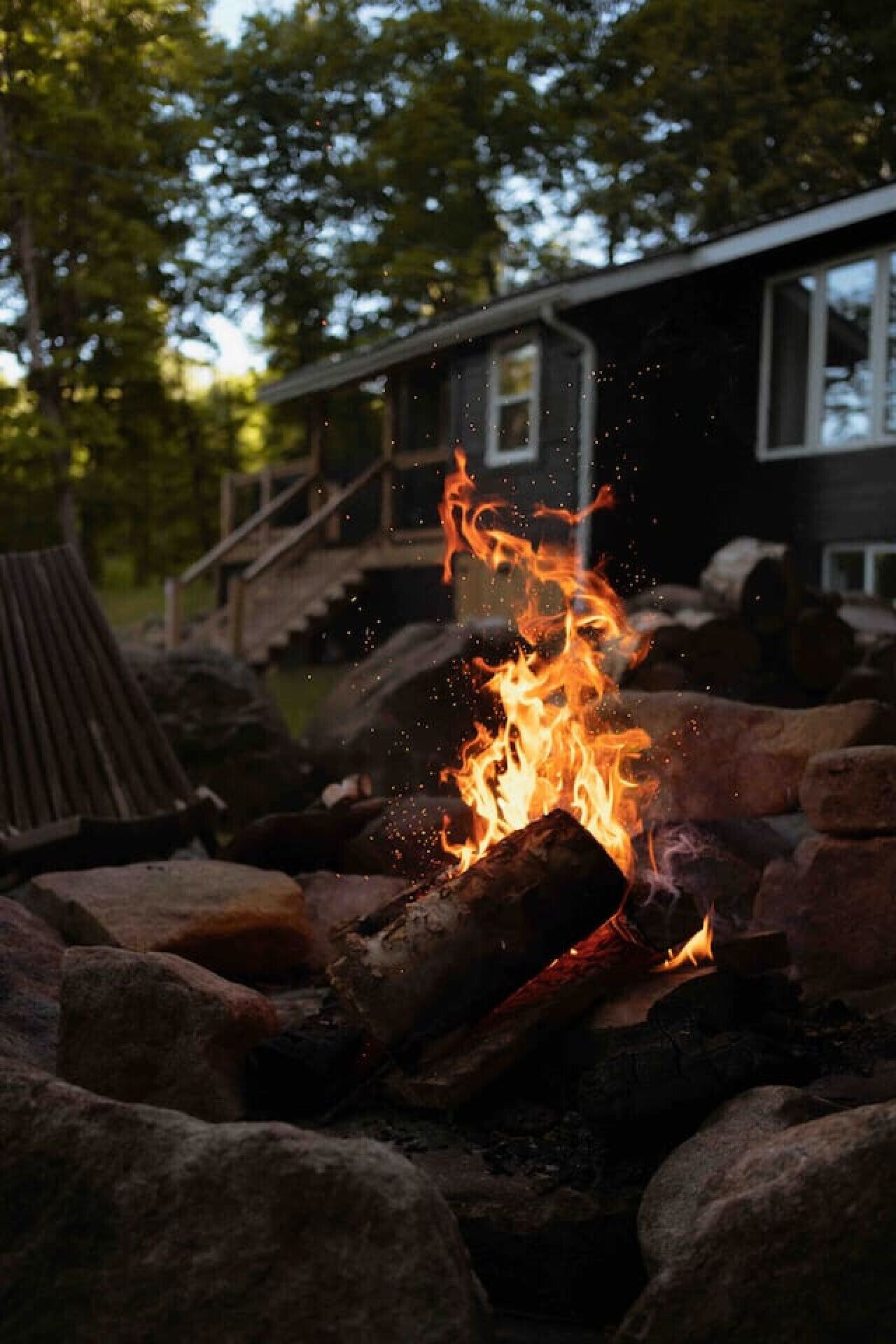 Ein Lagerfeuer mit Holzscheiten und Steinen, umgeben von einer Holzterrasse und einem Haus mit Treppe.