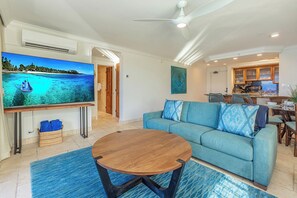 A living room with a large flat-screen TV, a blue sofa, a wooden coffee table, and a kitchen area in the background. - Oceanfront corner unit w/ panoramic views in Kahana (Lahaina)