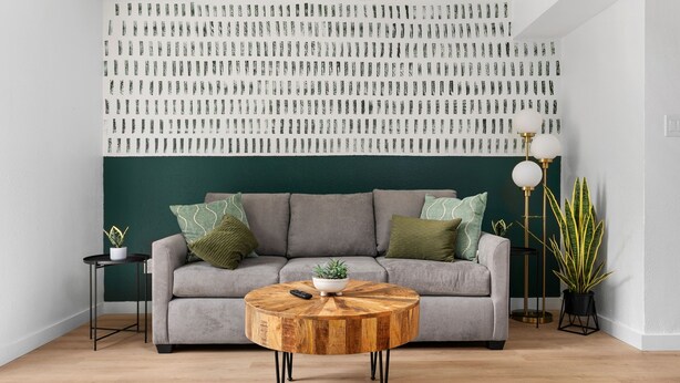 A living room with a round wooden coffee table, a grey sofa with green cushions, a wall with a patterned design, and a floor lamp. - Southport Combined (Austin)