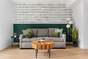 A living room with a round wooden coffee table, a grey sofa with green cushions, a wall with a patterned design, and a floor lamp.