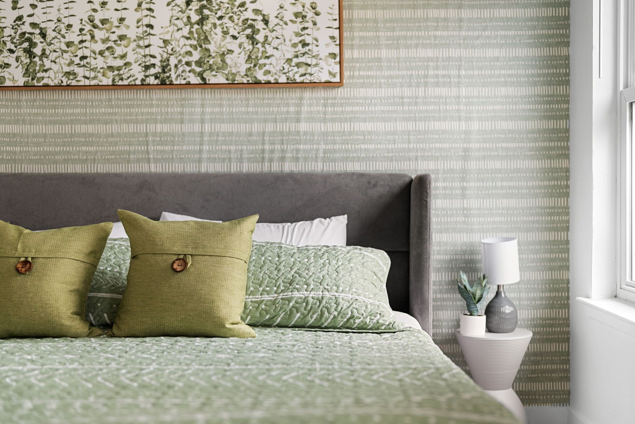 Een netjes opgemaakt bed met groen beddengoed en kussens, een grijs hoofdbord, en een nachtkastje met een lamp en een kleine plant.