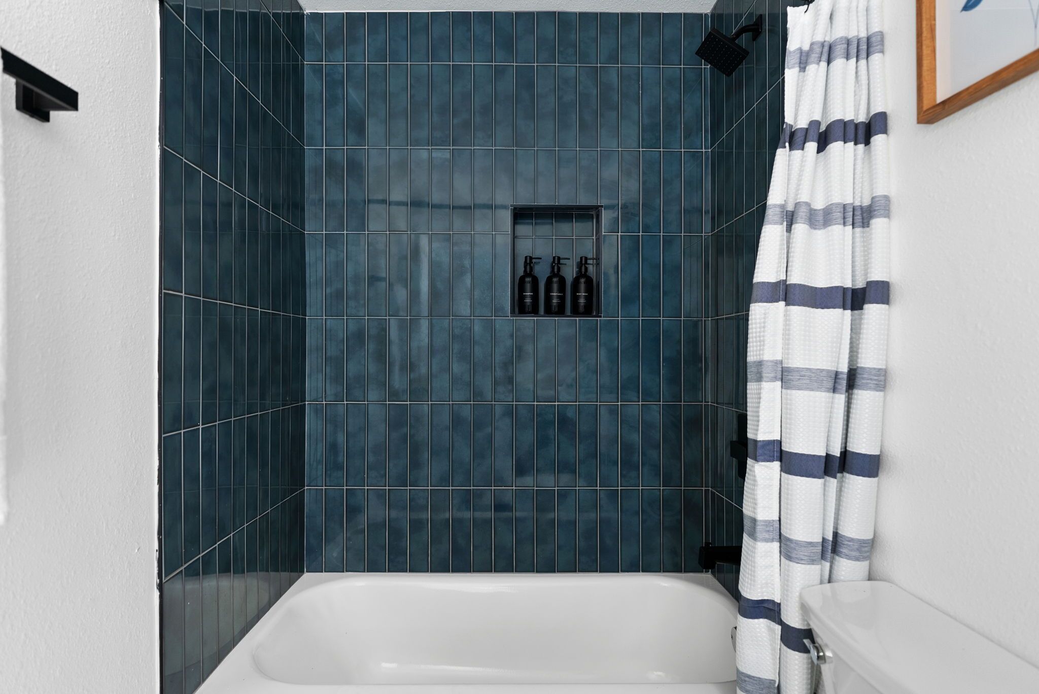 A bathroom with a bathtub, dark blue tiled walls, a framed picture on the wall, and a shower curtain.