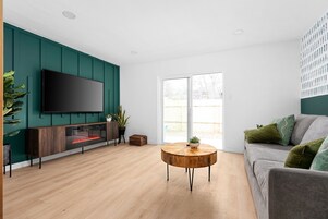 A modern living room with a flat-screen TV mounted on a teal wall, a round wooden coffee table, and a grey sofa with green cushions.