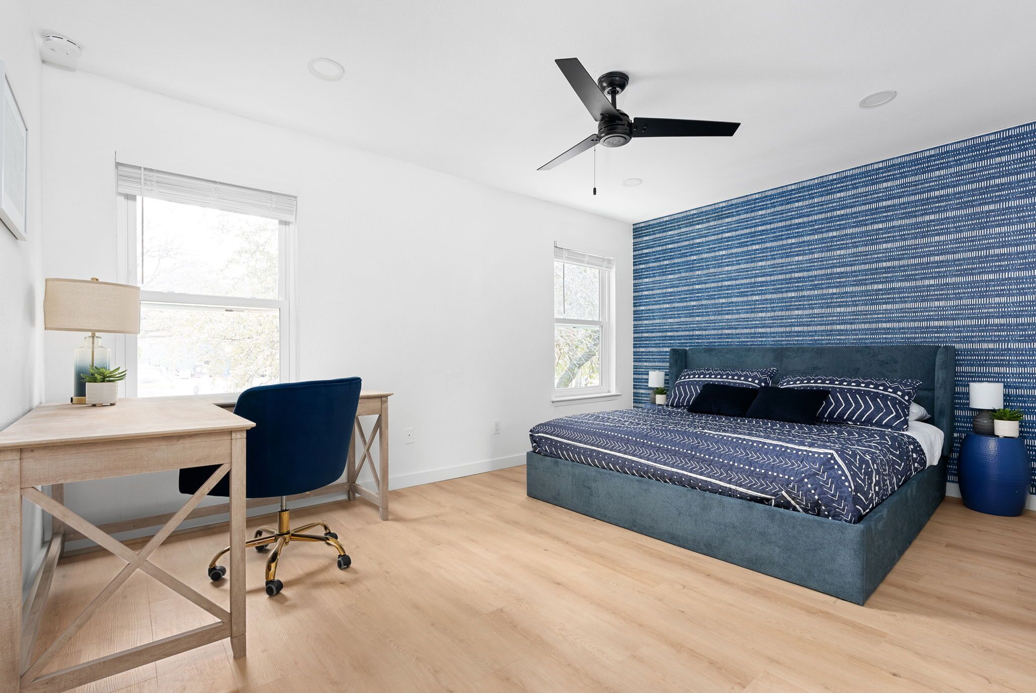 Une chambre à coucher comprenant un lit, un bureau, une chaise, un ventilateur de plafond et de grandes fenêtres.