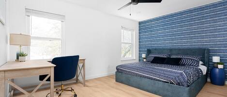 Une chambre à coucher comprenant un lit, un bureau, une chaise, un ventilateur de plafond et de grandes fenêtres.