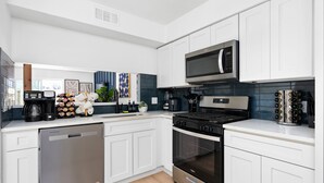 A modern kitchen with white cabinetry, a stainless steel dishwasher, a built-in oven, a microwave, and a coffee maker. - Southport Combined (Austin)