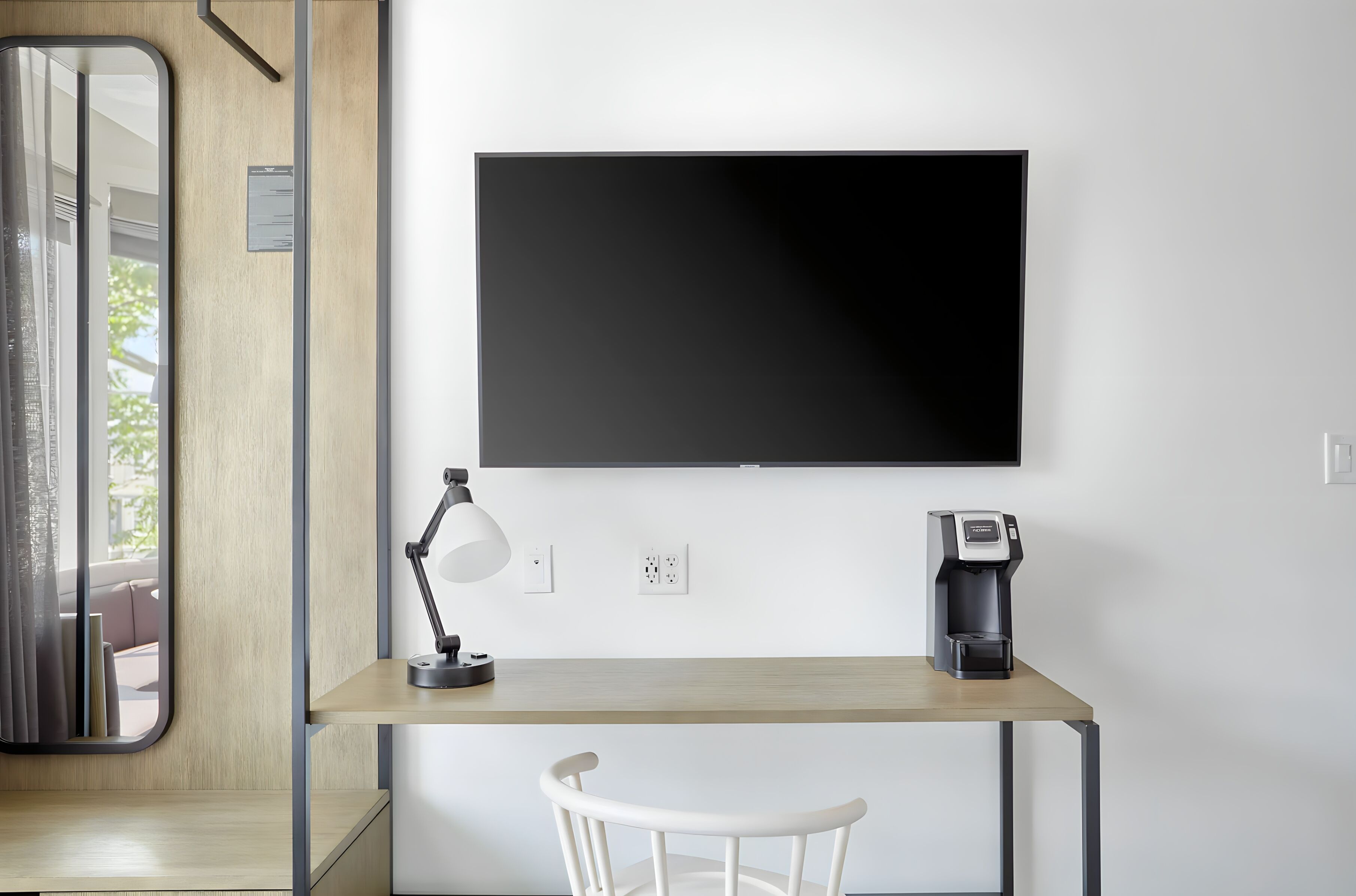 Un espace de travail moderne comprenant un bureau, une chaise, une machine à café, une lampe et un grand écran noir fixé au mur.