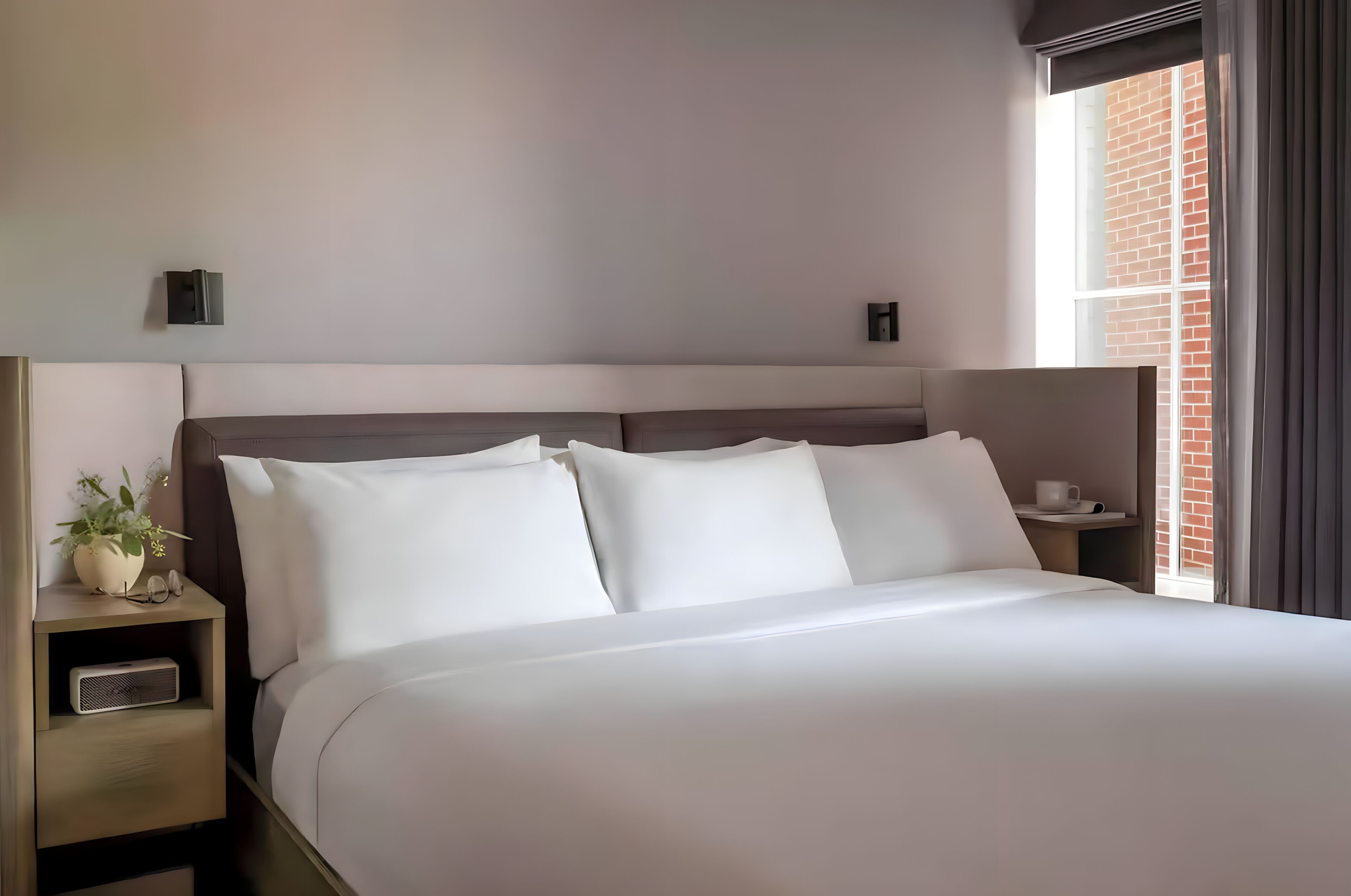Une chambre d’hôtel avec un grand lit, une table de chevet agrémentée d’une plante et une fenêtre donnant sur un immeuble en briques.