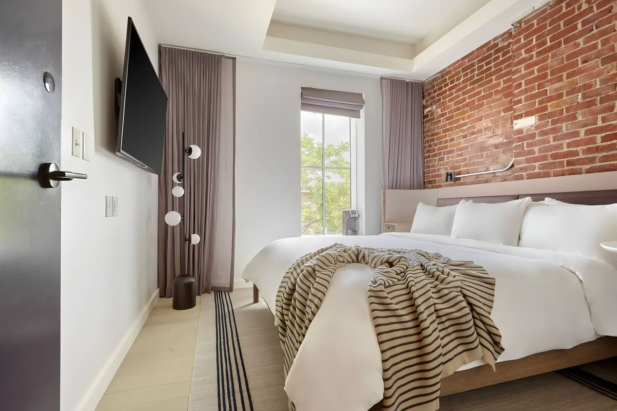 A modern hotel room with a large bed, a brick accent wall, a flat-screen TV, and a view of the outdoors.