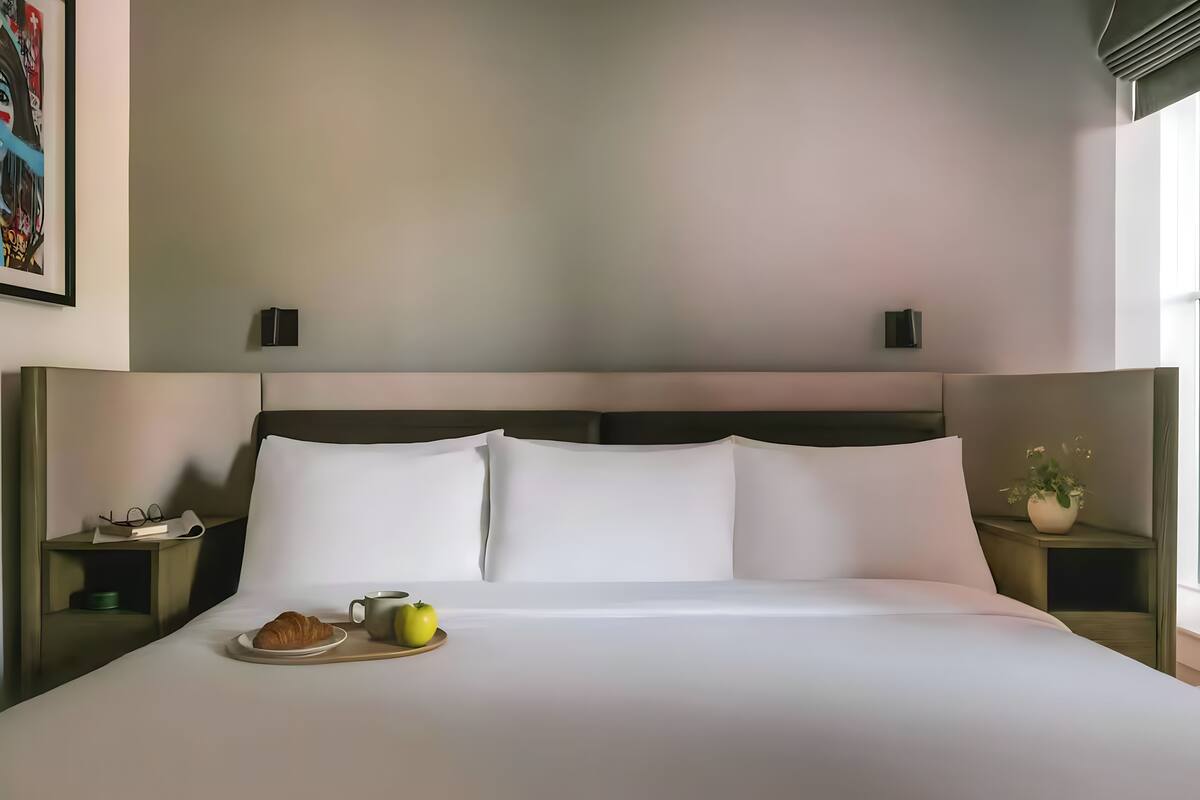 A neatly made bed with white linens, a bedside table with a book and glasses, and a small potted plant on the nightstand.
