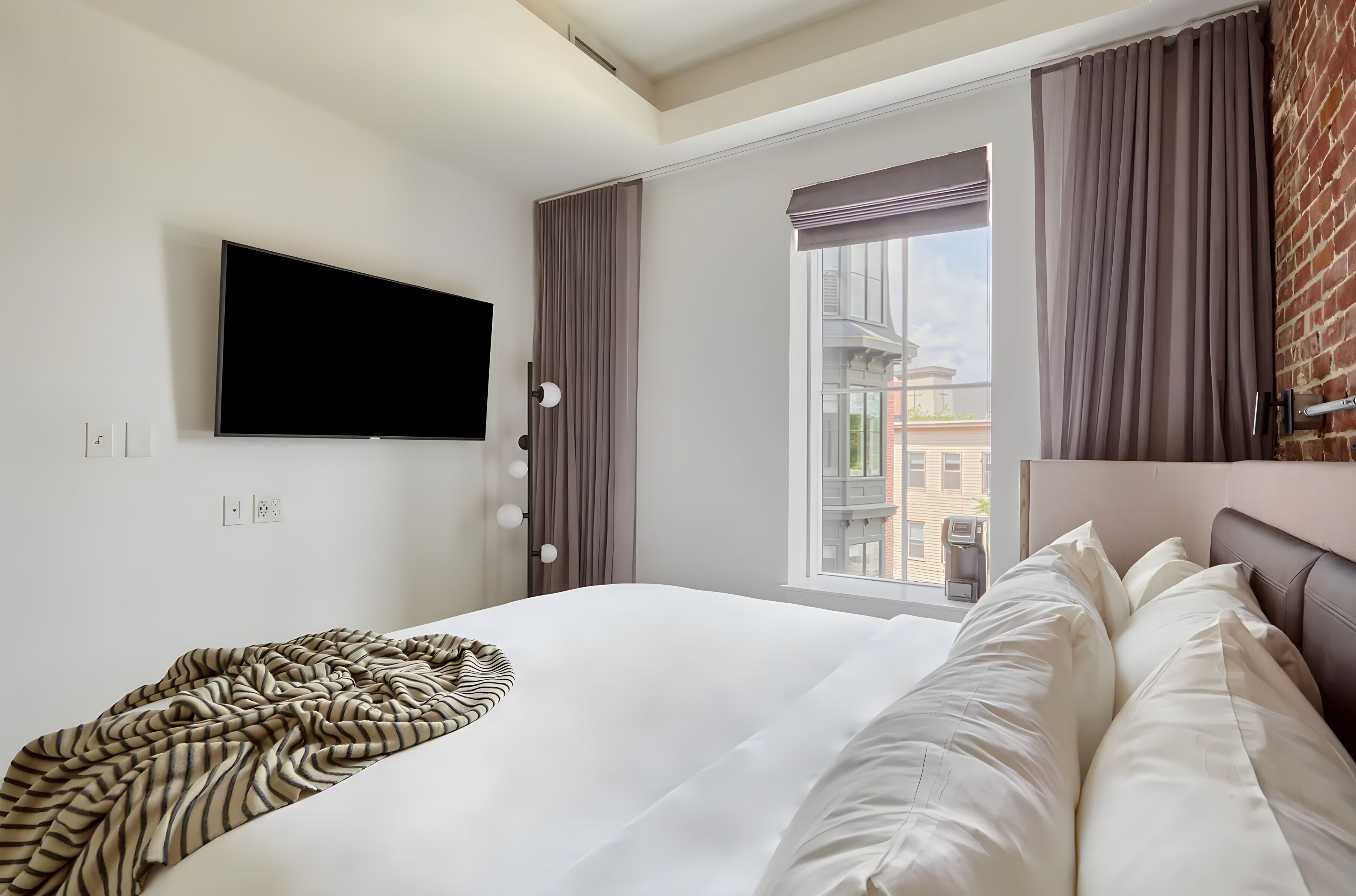 A hotel room with a large bed, a flat-screen TV mounted on the wall, a window with curtains, and a brick wall.
