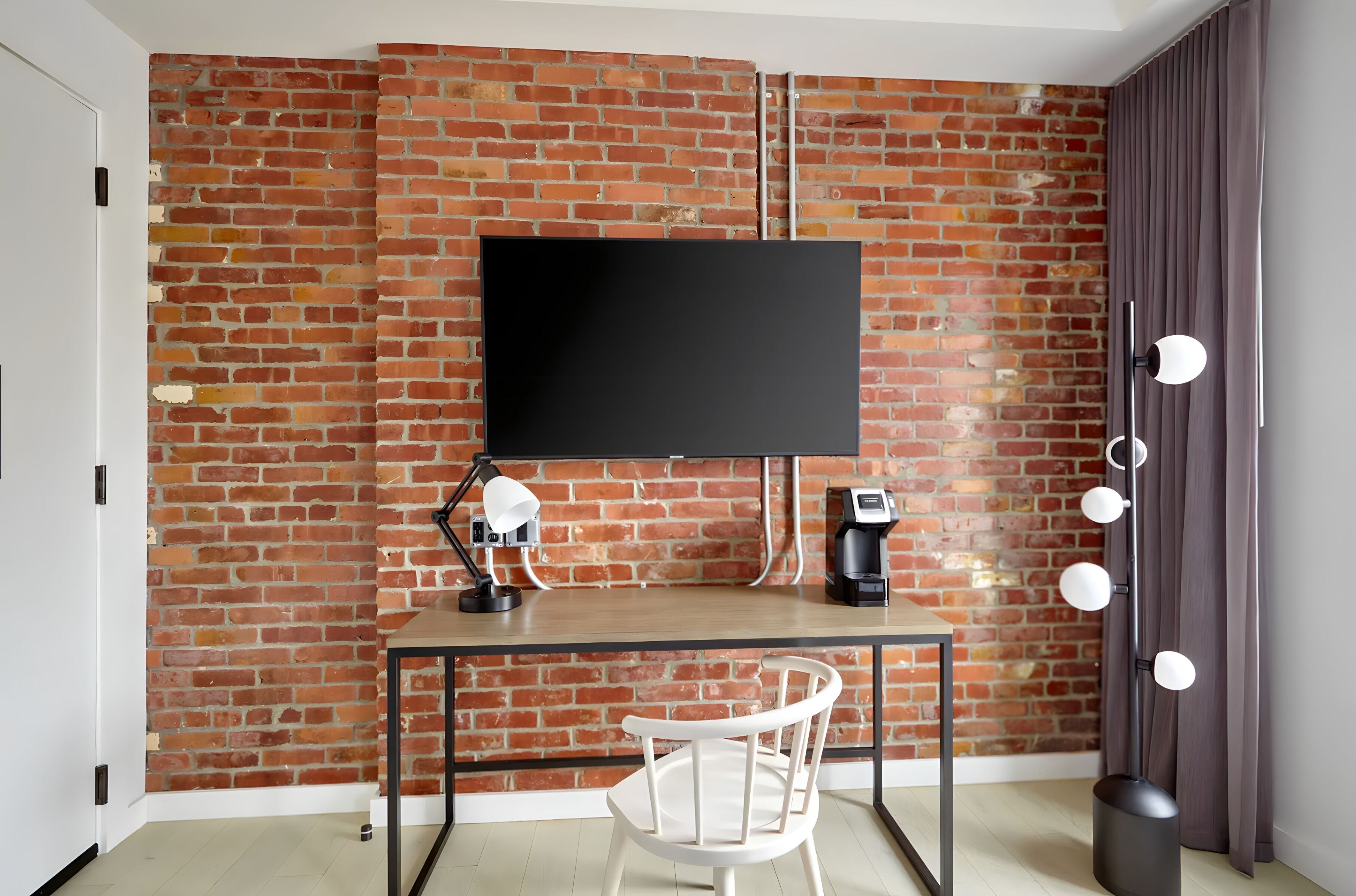 A room with a brick wall, a flat-screen TV mounted above a desk, a white chair, a lamp, a coffee machine, and a floor lamp with multiple bulbs.