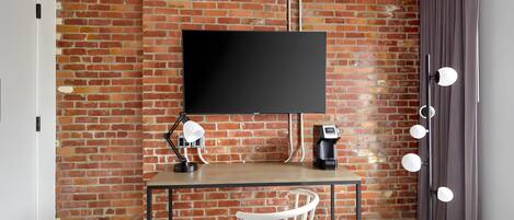 A room with a brick wall, a flat-screen TV mounted above a desk, a white chair, a lamp, a coffee machine, and a floor lamp with multiple bulbs.