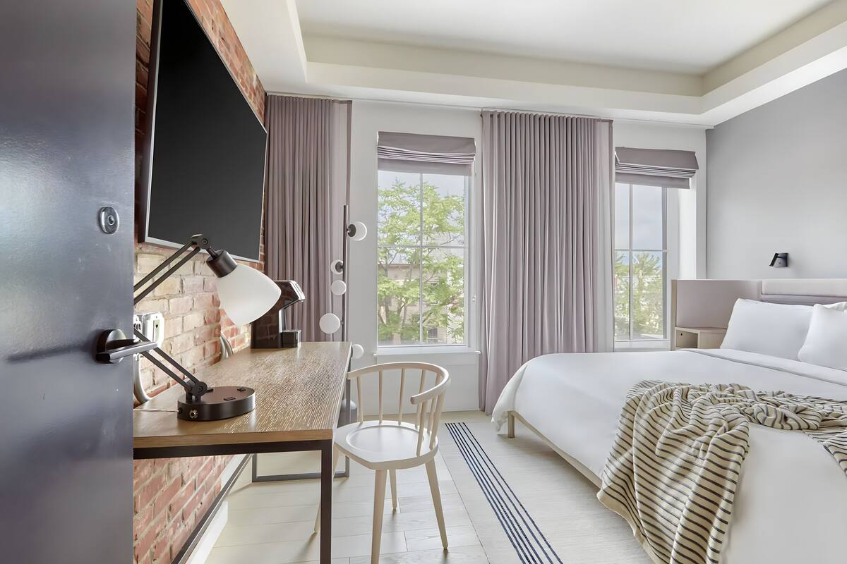 A hotel room with a bed, a desk with a lamp, a chair, and a window with curtains.