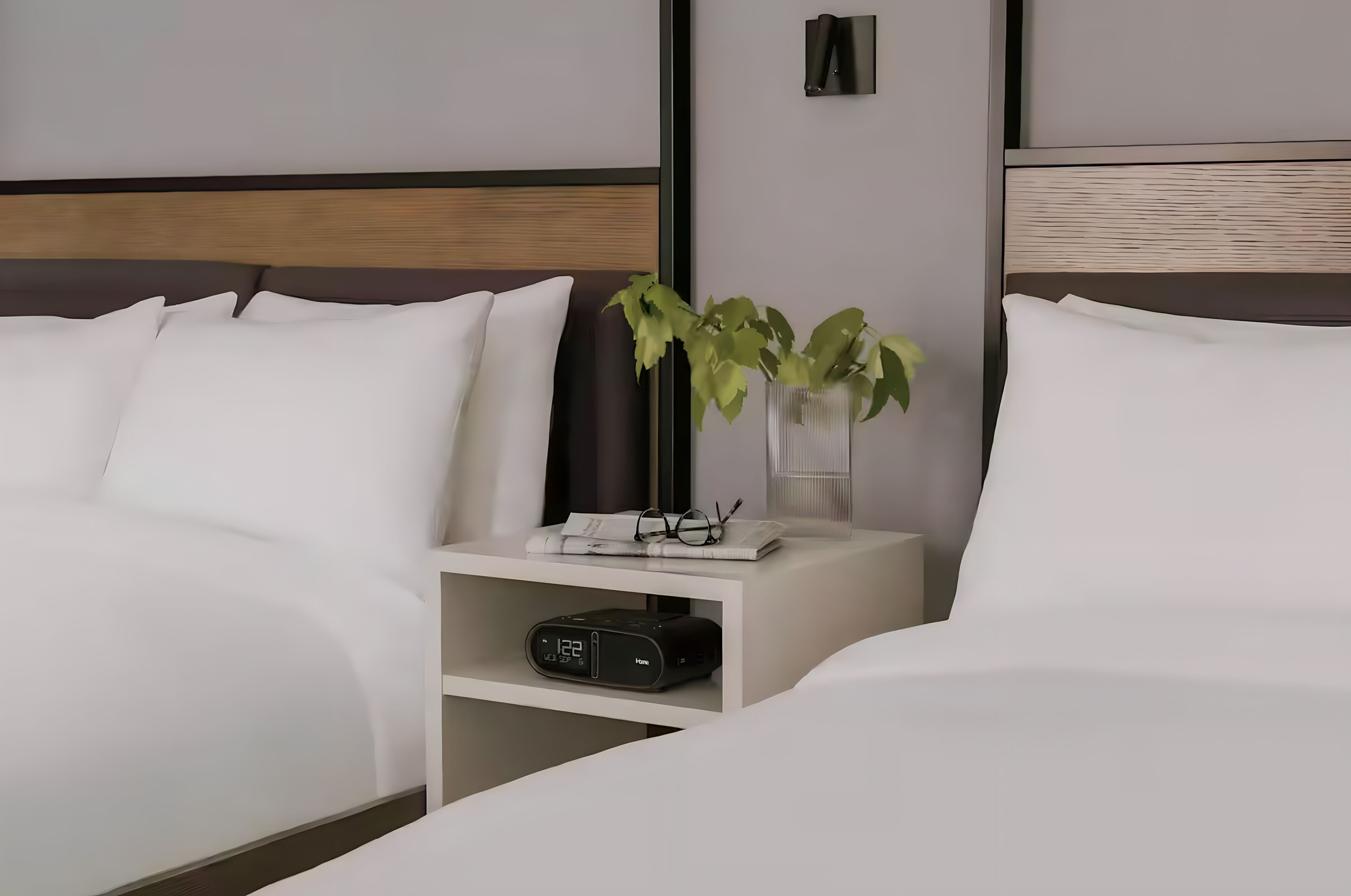 Un lit bien fait, avec une literie blanche, une table de chevet sur laquelle on peut voir une horloge, des lunettes et une plante.