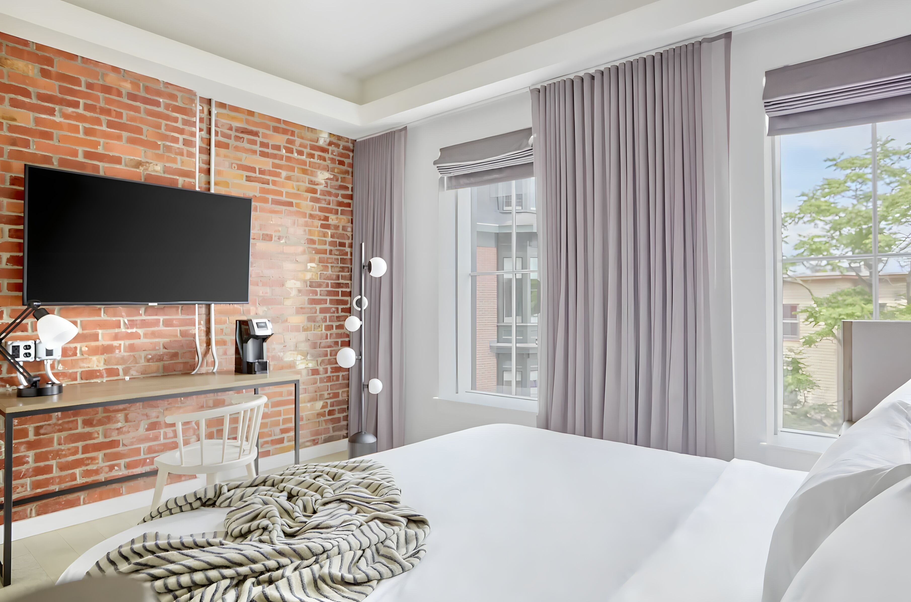 Habitación de hotel con pared de ladrillo, televisor de pantalla plana, escritorio con silla, cama con ropa blanca y ventana con cortinas.