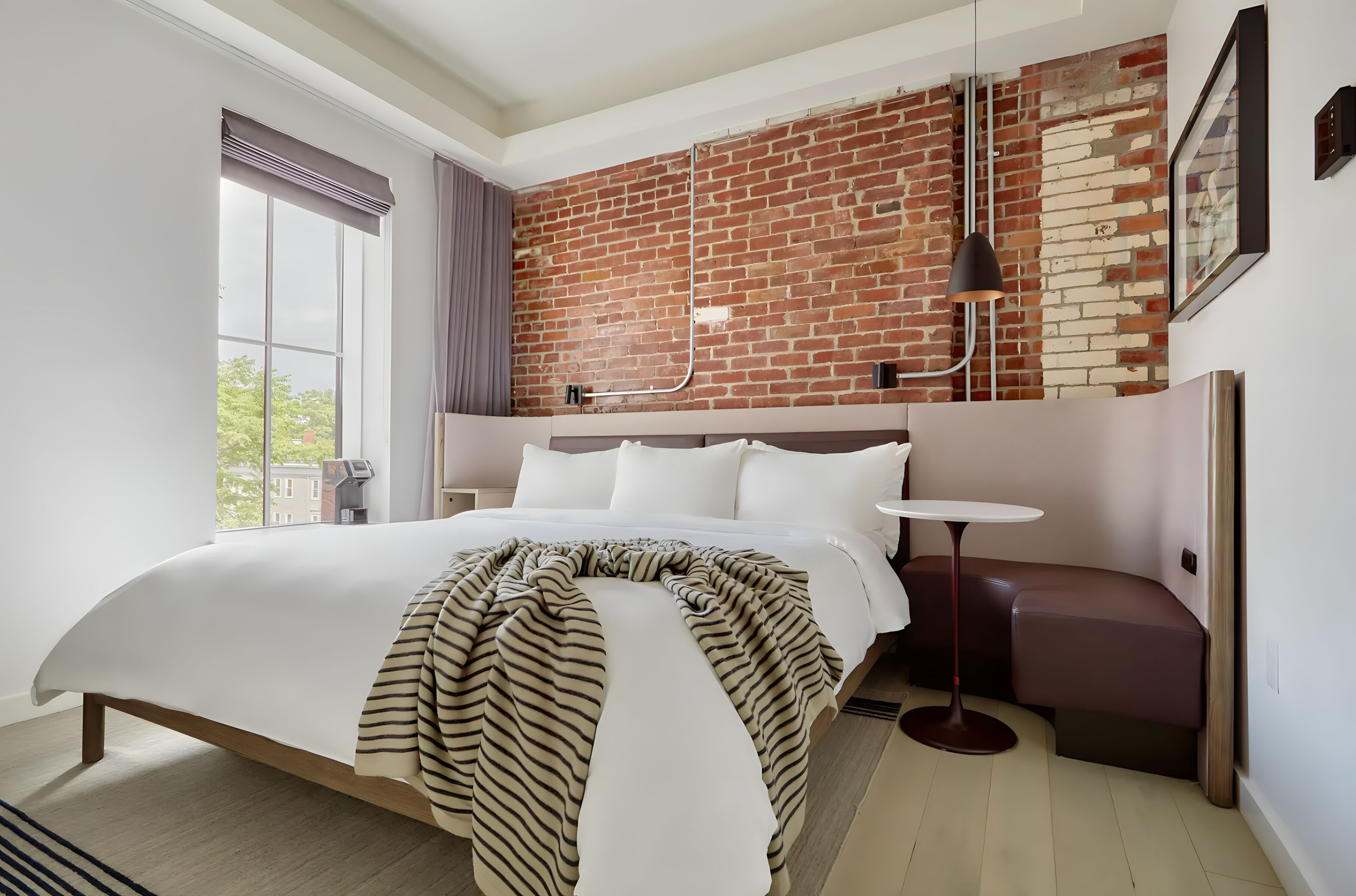 A modern hotel room with a large bed, a small round table, a bench, and a brick wall.
