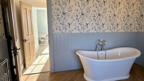 Un baño con una bañera independiente, papel tapiz azul claro con motivos florales y suelo de madera.