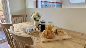 Una mesa de madera con un florero de flores blancas, una cafetera, una jarra de jugo de naranja y croissants.