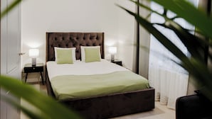 Une chambre à coucher avec une tête de lit capitonnée, deux tables de chevet avec des lampes, une couvre-lit verte et une vue à travers des rideaux transparents.