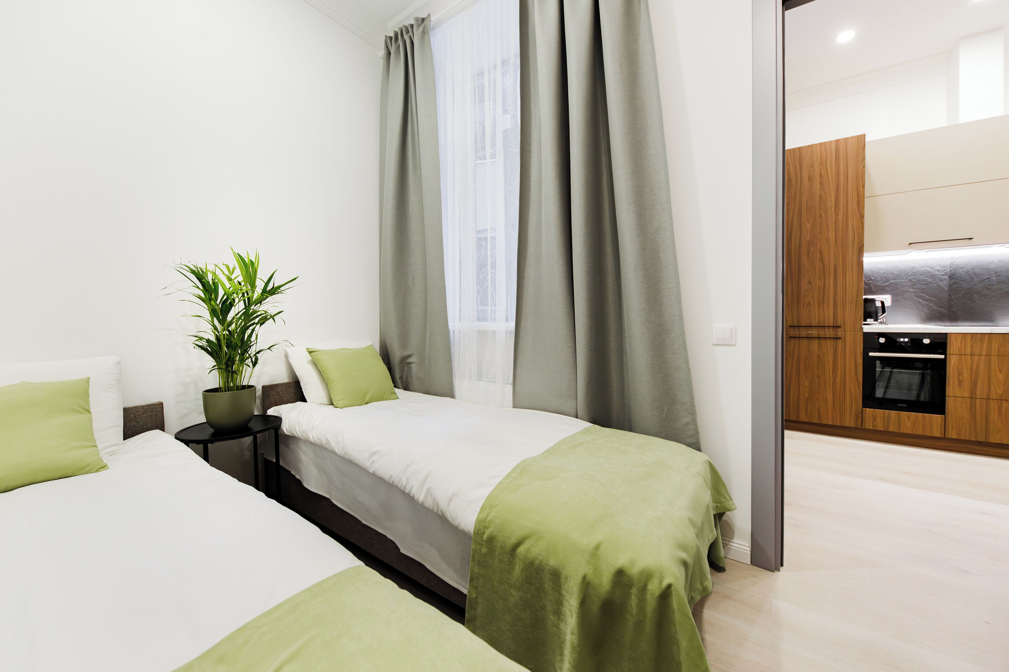 A modern hotel room with two beds, a small table with a plant, and a view of the kitchen.