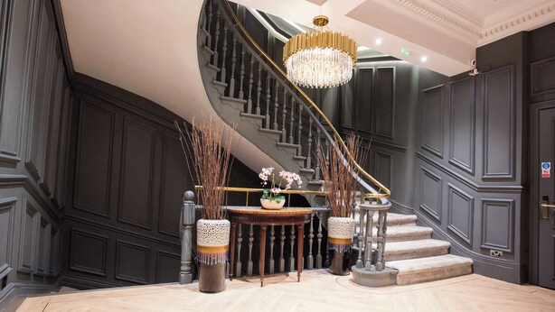 Eine imposante Treppe mit einer Chandelüster, dunklen Paneelen und einem runden Tisch mit Zierpflanzen.