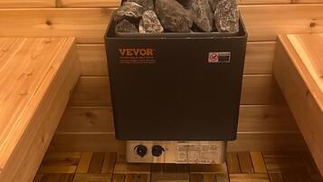 Una sauna con una stufa nera recante l'etichetta 'VEVOR' e un'avvertenza. Il pavimento è in legno con un motivo a spina di pesce.