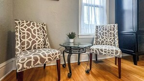Two patterned chairs, a small glass table with a plant, and a dark wooden cabinet in a room with wooden flooring and a window with sheer curtains. - Enjoy the entire main level of this Historic Victorian duplex form the 1800s. Located in the heart of Cambridge. Unwind in private 4-person sauna, king size beds, quaint front porch and fully stocked kitchen. Walk to everything in town. (Cambridge)