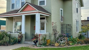 A two-story house with a porch, a front door, and a small garden area with plants and a bicycle. - Enjoy the entire main level of this Historic Victorian duplex form the 1800s. Located in the heart of Cambridge. Unwind in private 4-person sauna, king size beds, quaint front porch and fully stocked kitchen. Walk to everything in town. (Cambridge)