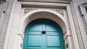 Une porte à deux battants, de couleur verte, dotée d’une poignée circulaire, encadrée par un arc en pierre.