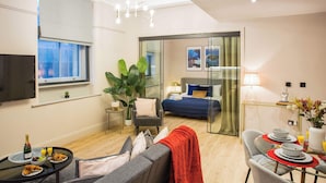 Une chambre d’hôtel moderne comprenant un lit, un coin salon avec un canapé et un fauteuil, une table à manger avec des assiettes et des verres, et un téléviseur fixé au mur.
