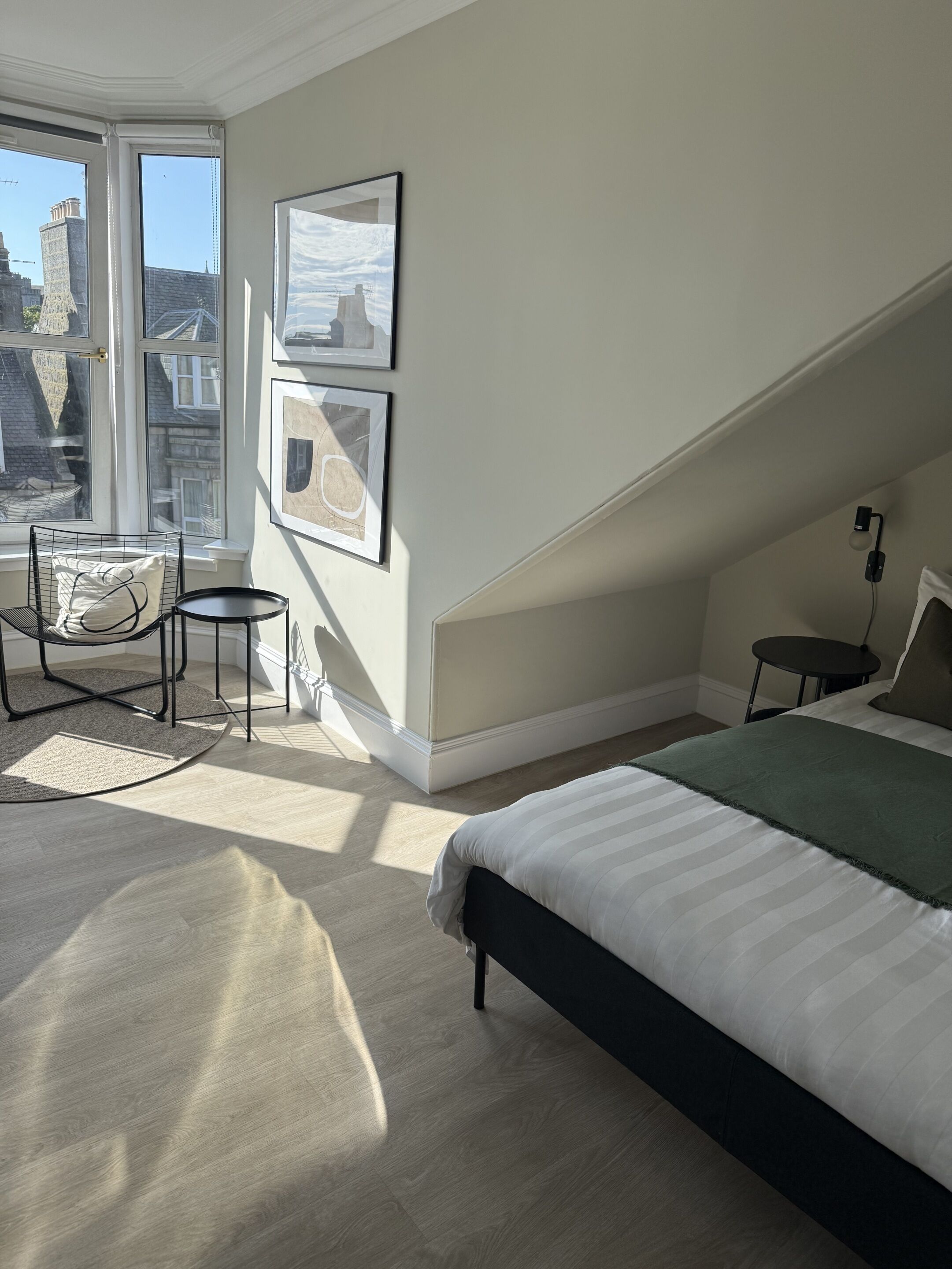 A bedroom with a bed, a chair, a small table, and a window with a view of buildings.