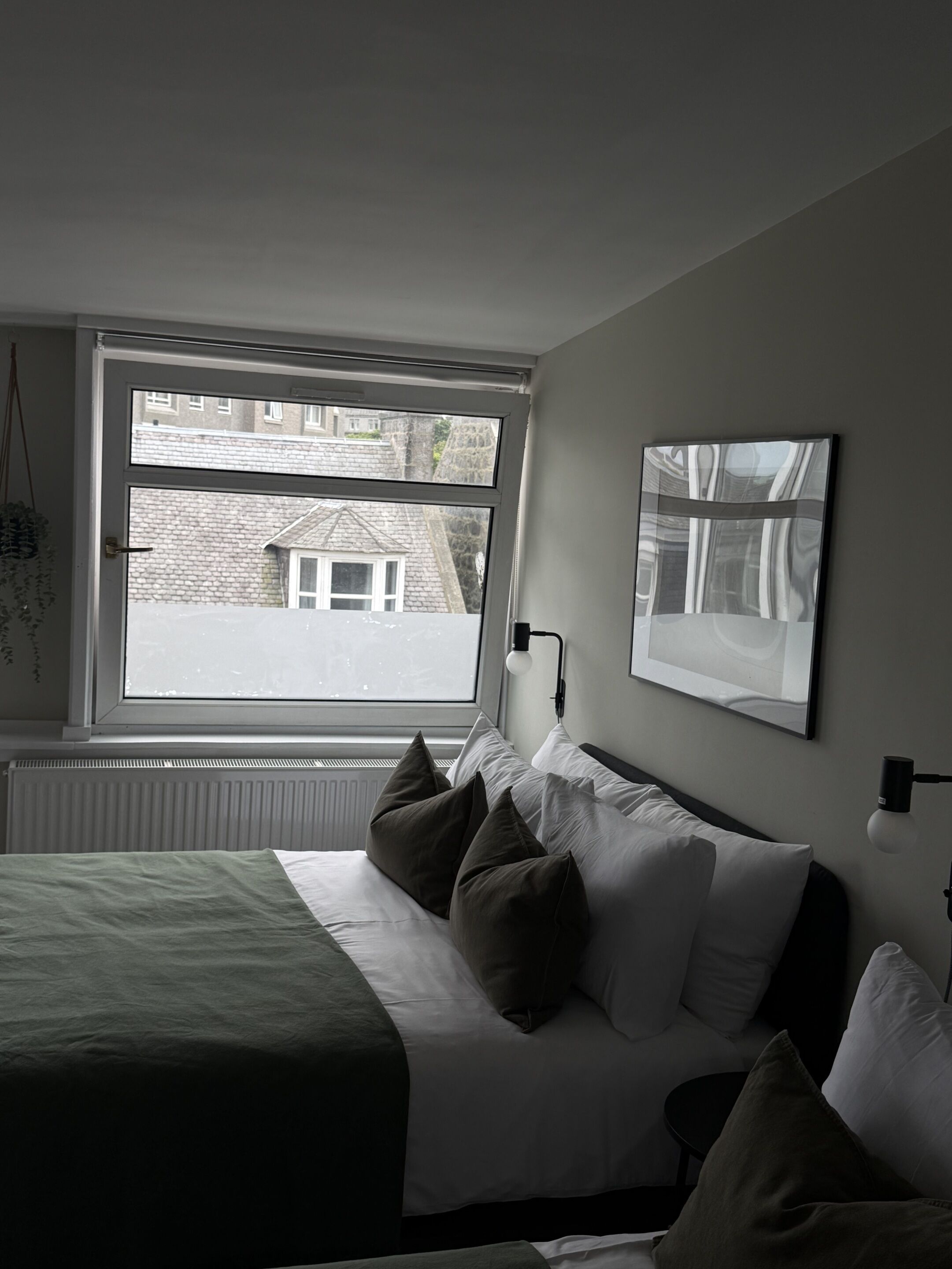 A bedroom with a large window, a bed with multiple pillows, a radiator, and two wall-mounted light fixtures.