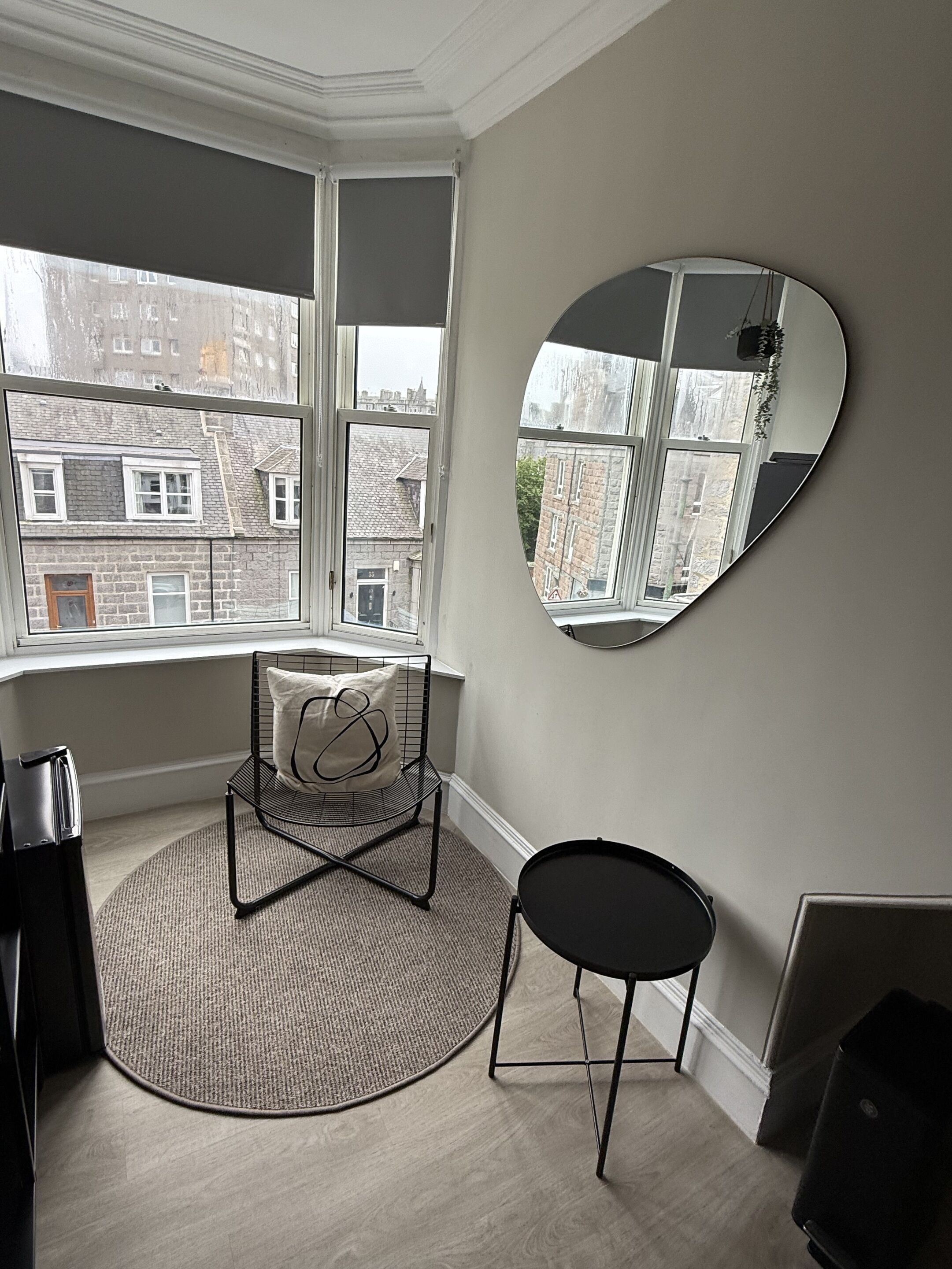 A room with a large window, a round mirror, a black chair, a small table, and a rug.