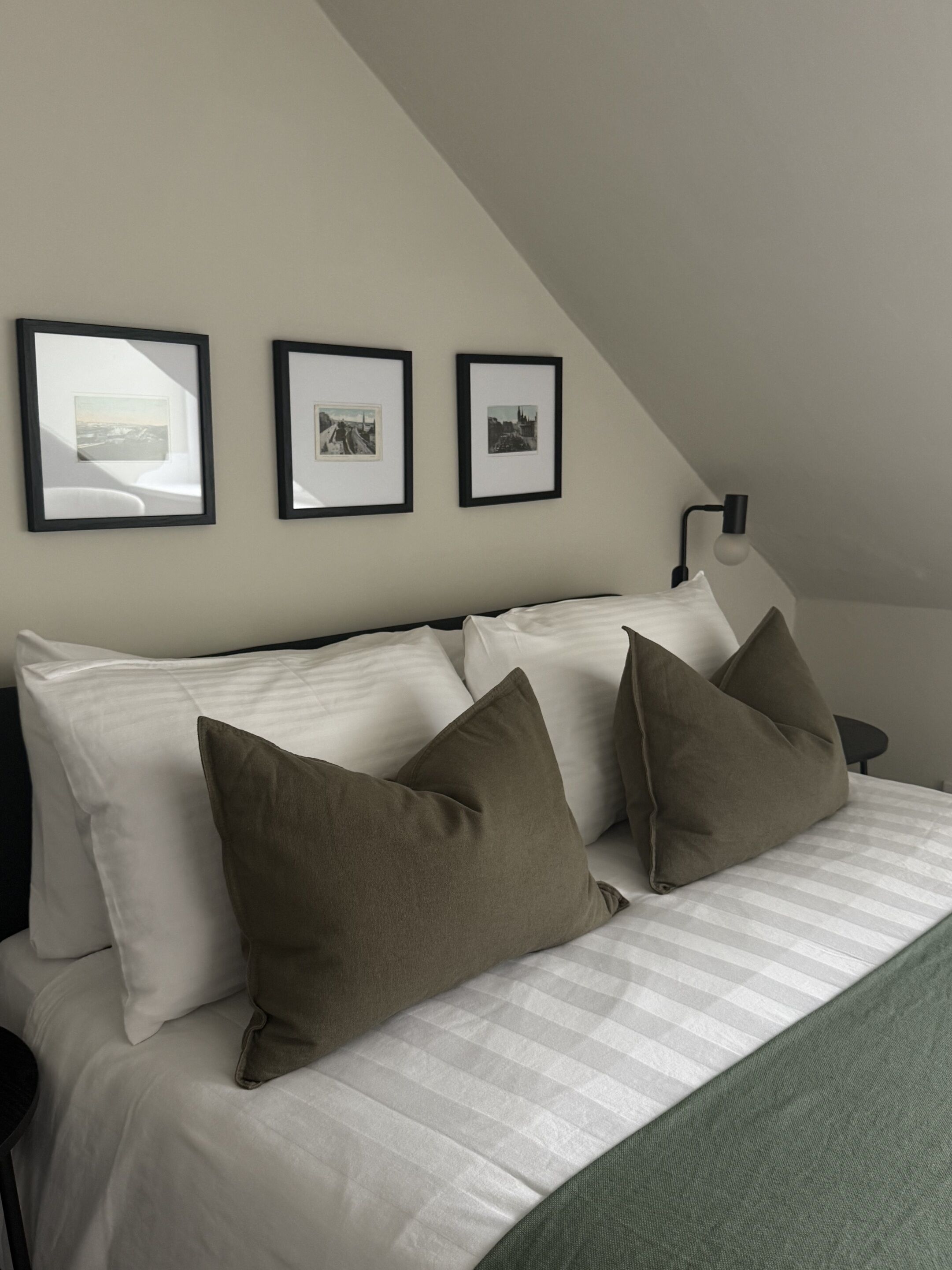 Een netjes opgemaakt bed met kussens, een lamp en drie ingekaderde foto’s aan de muur.