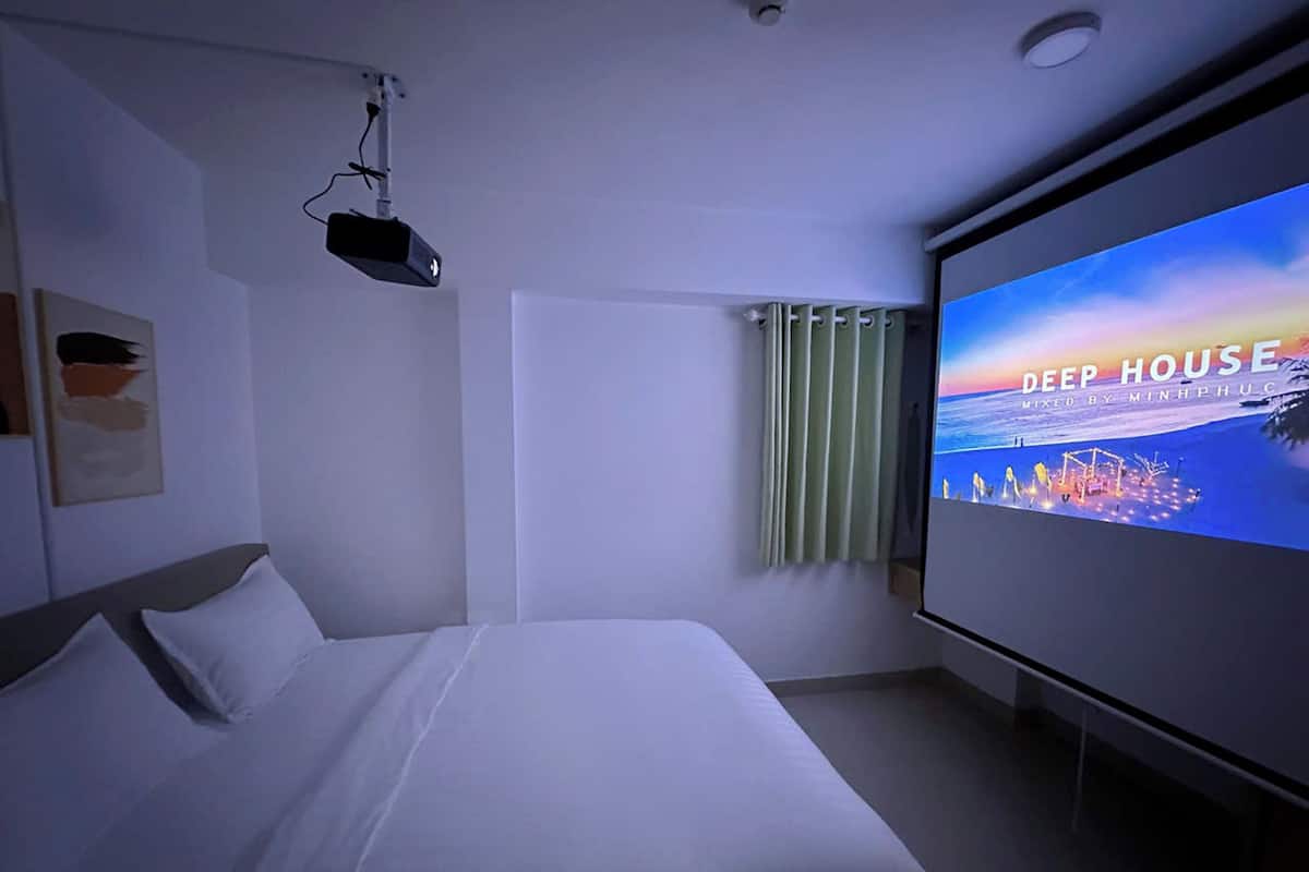 Hotelkamer met een bed, een scherm waarop een 'DEEP HOUSE' muziekvideo wordt getoond, een raam met gordijnen en een aan het plafond gemonteerde projector.