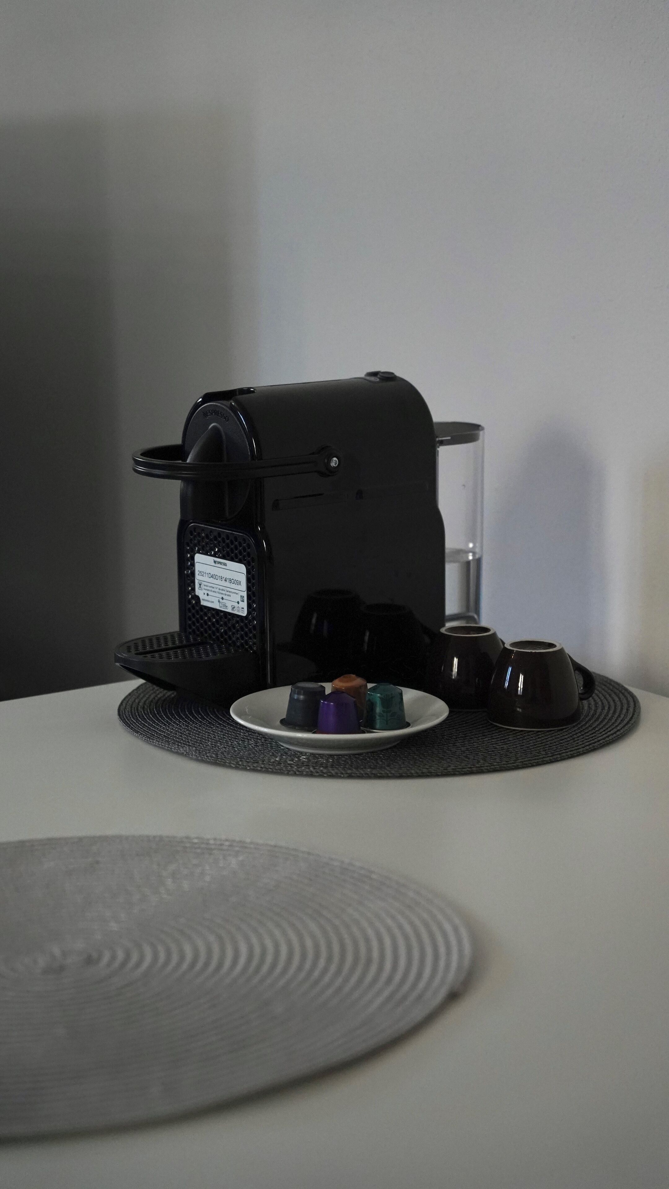 En kaffemaskin med en uppsamlingsbricka, ett glas vatten och en tallrik med kaffekoppar och en kaffekapsel.