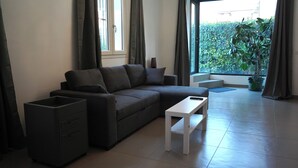 Un salon moderne avec un canapé gris, une petite table blanche et un meuble noir. On y trouve une fenêtre avec des rideaux, une plante en pot et une vue sur la verdure à l’extérieur.