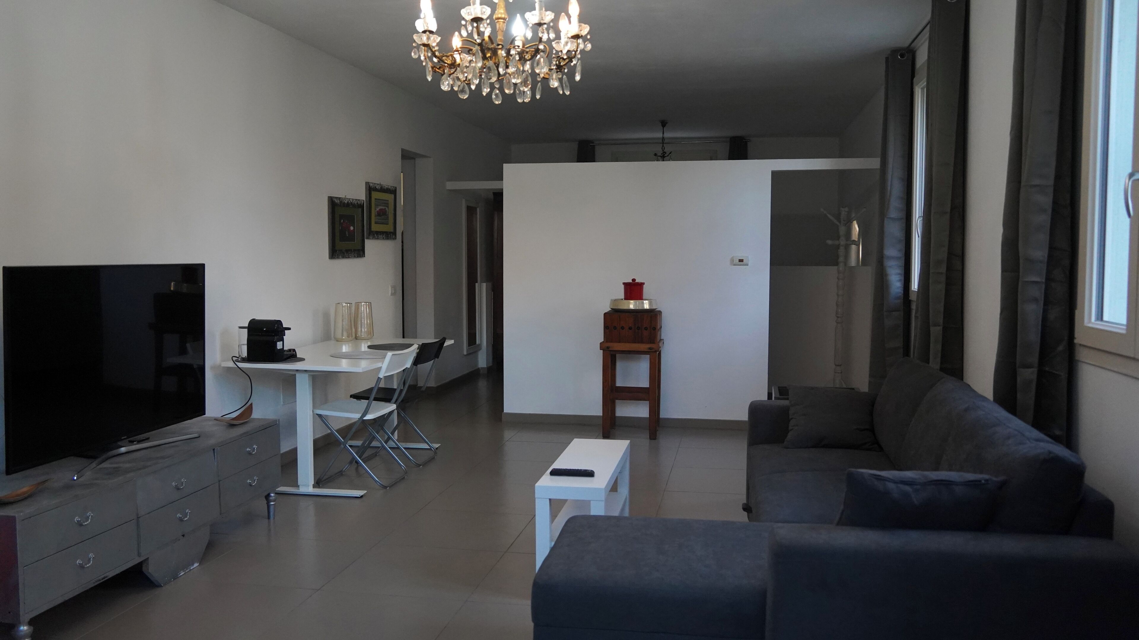 A modern living room with a flat-screen TV, a sofa, a dining table with chairs, and a chandelier.
