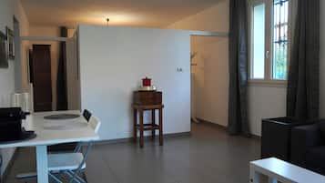 A modern living room with a white sofa, a dining table with chairs, a small wooden stool, and a window with curtains.