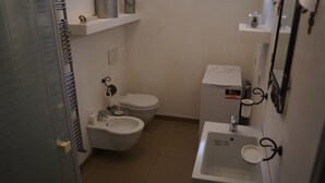 Une petite salle de bain comprenant des toilettes, un bidet et un lavabo. On y trouve une petite étagère avec une tasse et un récipient.