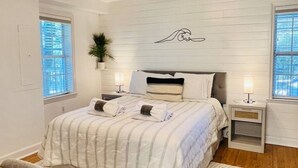 Ein Schlafzimmer mit einem Bett, Nachttischen, einem kleinen Tischchen, einer Zimmerpflanze und einem an der Wand hängenden Kunstwerk.
