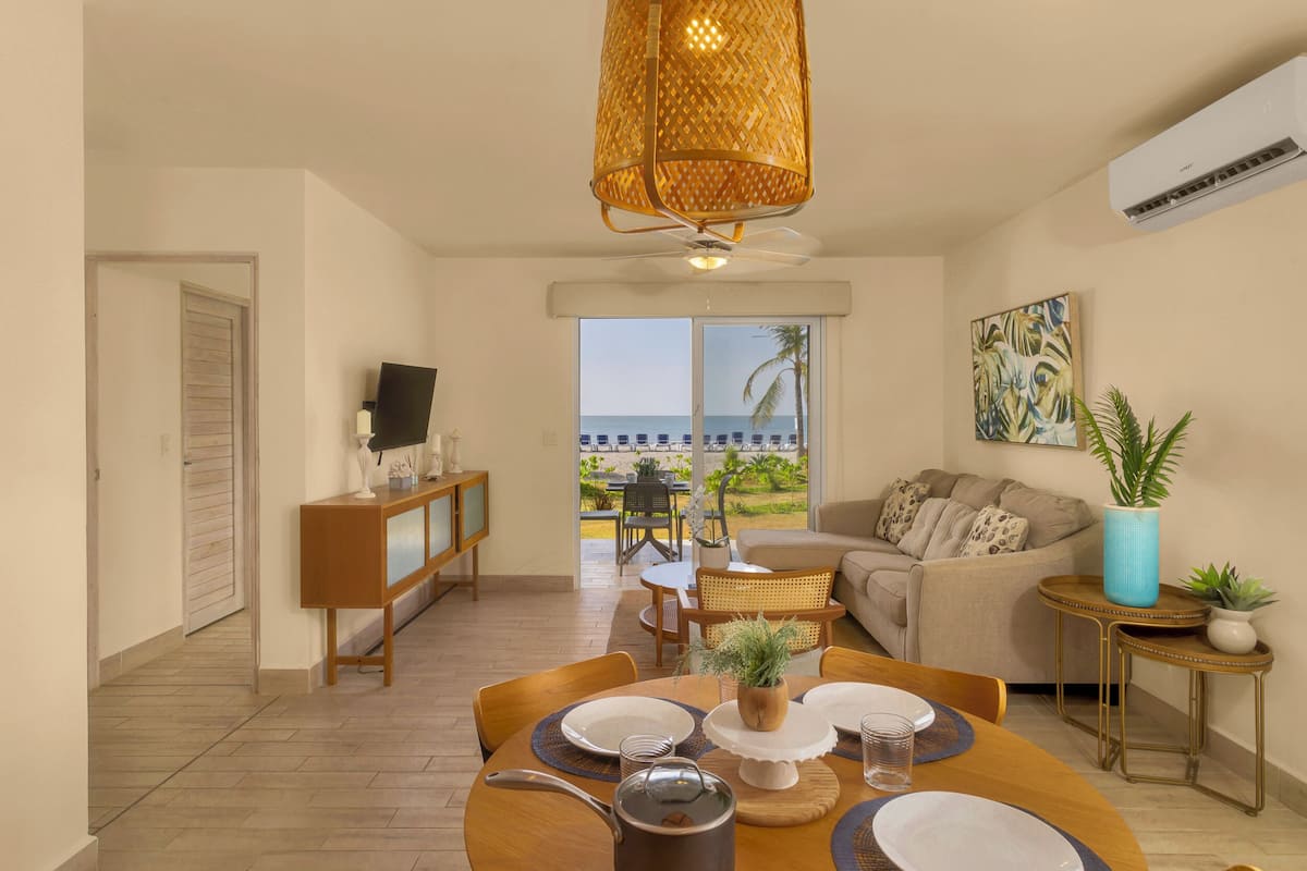 Una sala de estar con un sofá, una mesa de comedor preparada para cuatro personas, un televisor y un amplio ventanal con vistas al mar.