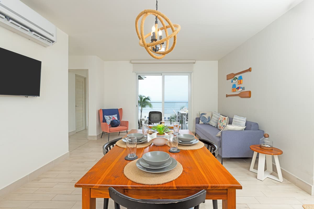 Un comedor con una mesa de madera dispuesta para cuatro personas, un televisor montado en la pared y vista al mar a través de un ventanal grande.