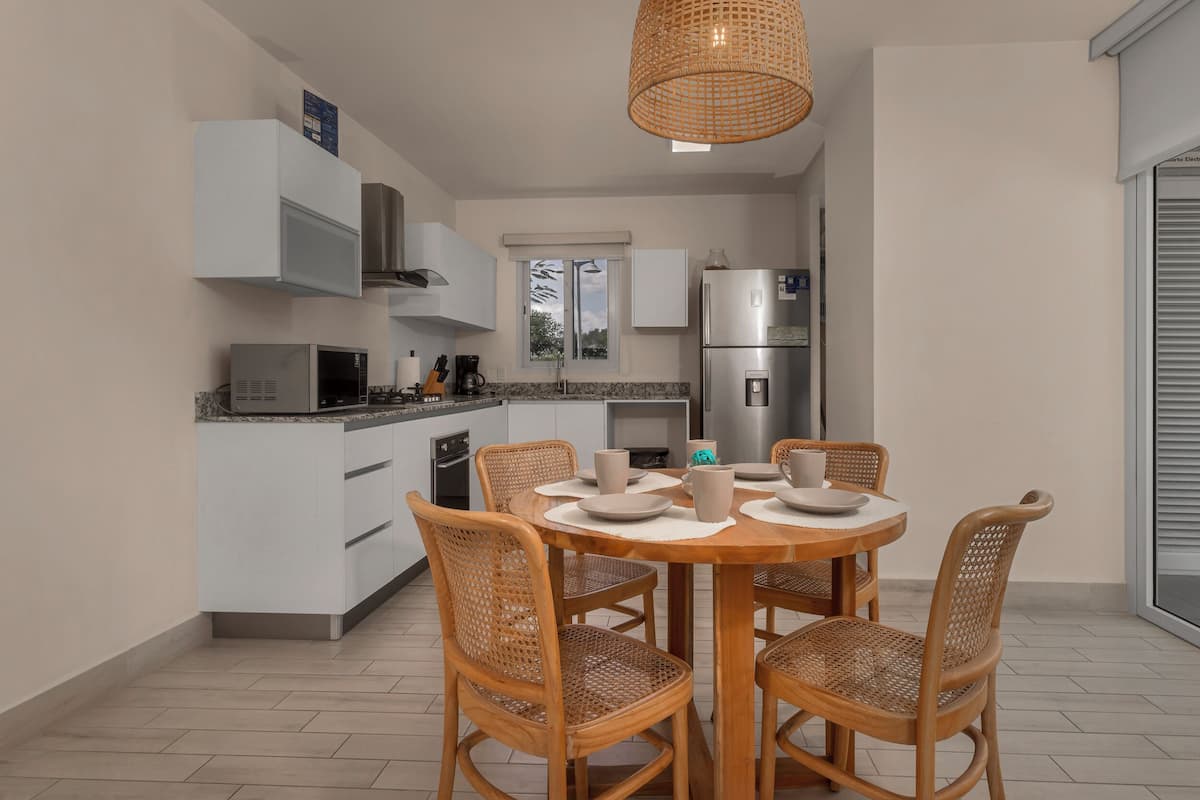 Una cocina moderna con armarios blancos, un refrigerador de acero inoxidable y un comedor con una mesa redonda y sillas de mimbre.