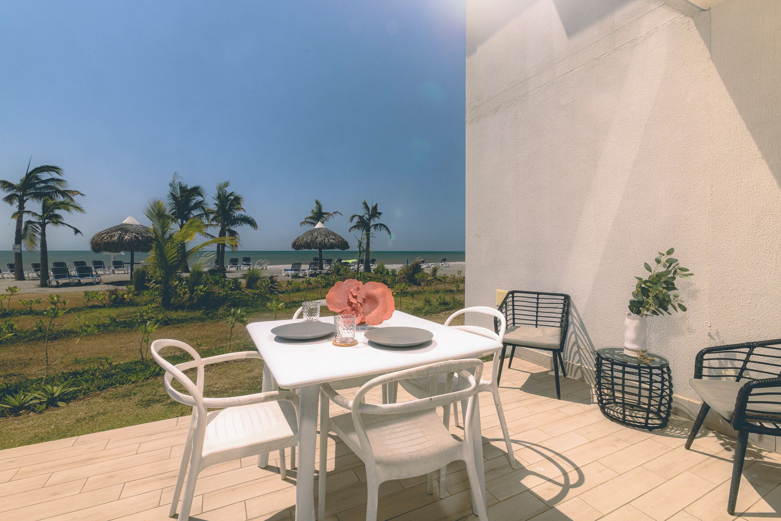Eine Terrasse mit weißem Tisch und Stühlen, einem Blumentopf und Blick auf den Strand mit Palmen und einem Reetdachschirm.