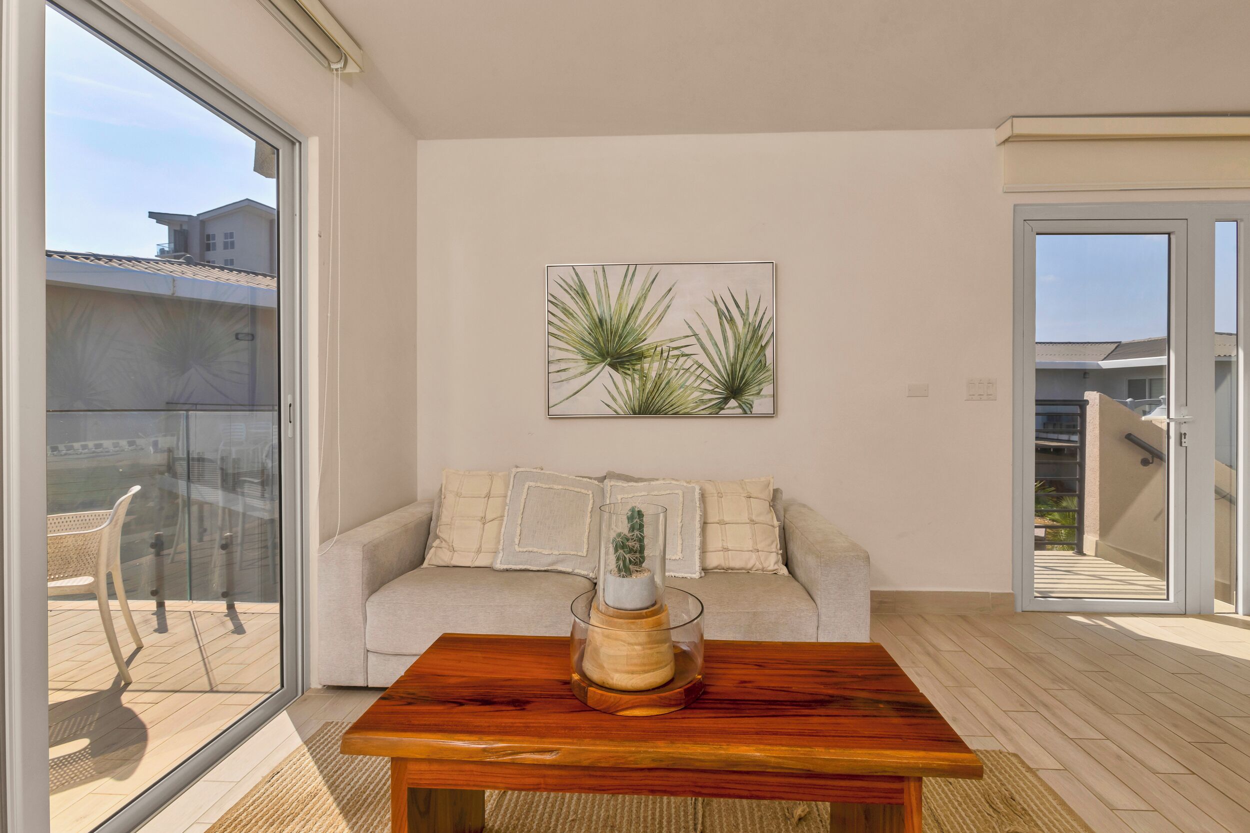 A modern living room with a glass sliding door, a wooden coffee table, a beige sofa with decorative pillows, and a framed plant artwork on the wall.