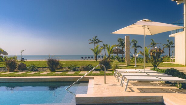 Zona de piscina con sillones reclinables, una palmera y vistas a la playa.