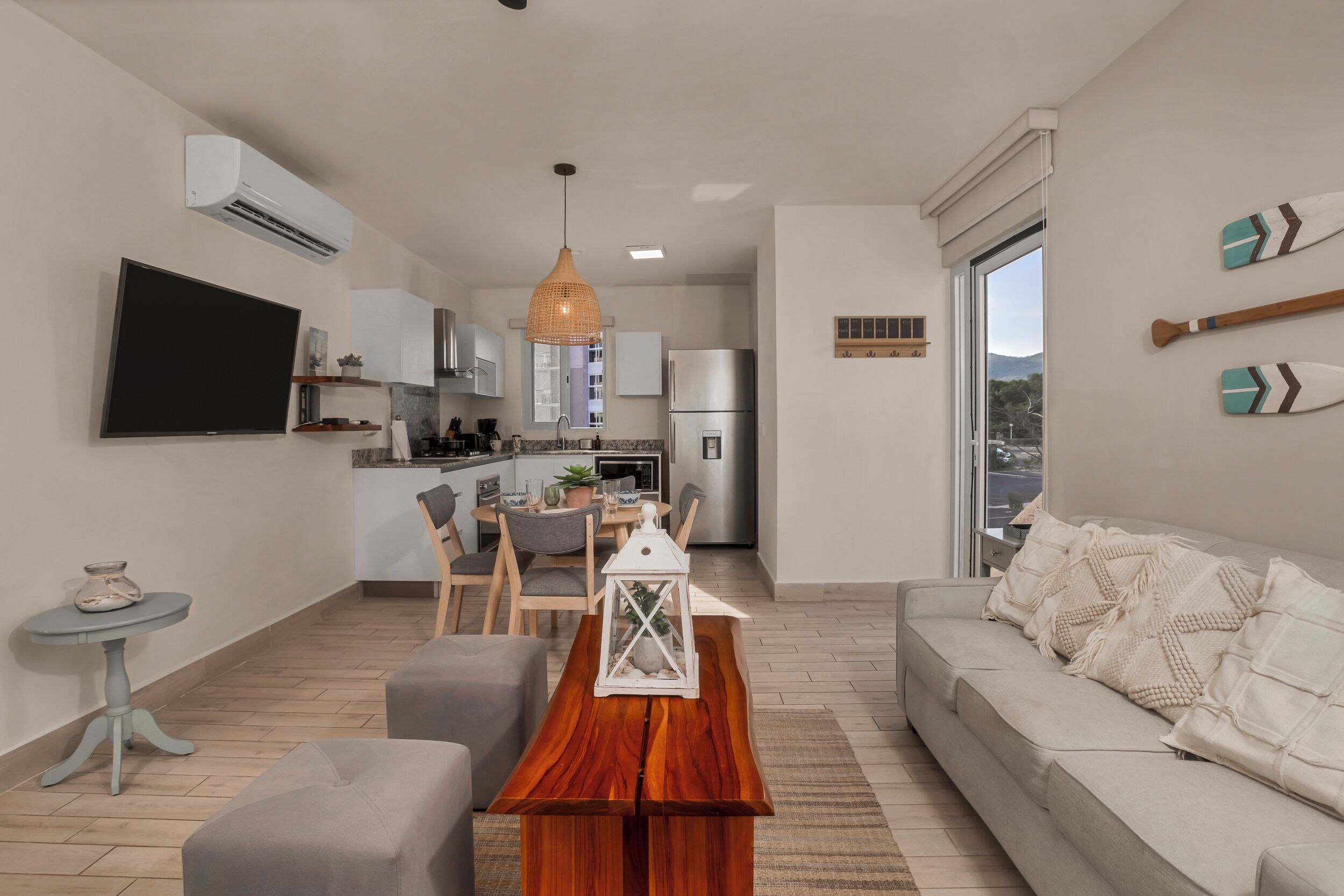 A modern living room with a sofa, a wooden coffee table, and a TV. There's a kitchen in the background with cabinets and a refrigerator.