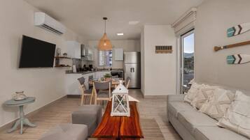 A modern living room with a sofa, a wooden coffee table, and a TV. There's a kitchen in the background with cabinets and a refrigerator.